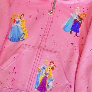 K&ouml;nnte beinhalten: Rosaer Kapuzenpullover mit Rei&szlig;verschluss und einem Spr&uuml;hfarben-Design mit Disney-Prinzessinnen. Zu den Prinzessinnen geh&ouml;ren Aschenputtel, Belle, Rapunzel, Elsa und Anna.