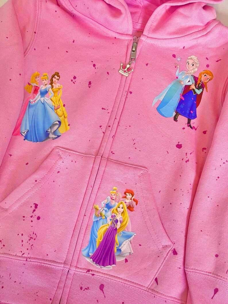 K&ouml;nnte beinhalten: Rosaer Kapuzenpullover mit Rei&szlig;verschluss und einem Spr&uuml;hfarben-Design mit Disney-Prinzessinnen. Zu den Prinzessinnen geh&ouml;ren Aschenputtel, Belle, Rapunzel, Elsa und Anna.