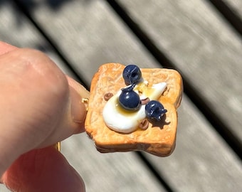 May include: A miniature toast ring. The ring features a slice of golden-brown toast topped with a dollop of white cream, blueberries, and a drizzle of syrup. A unique piece of food-themed jewellery.