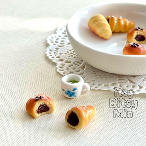 May include: Miniature pastries and a cup of tea are arranged on a white surface. The pastries include croissants and filled buns. A small white teacup with blue floral details holds green tea. The image includes the text "itsy Bitsy Min".