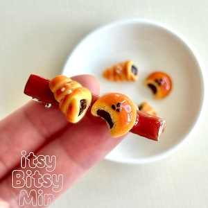 May include: A red hair clip adorned with miniature, detailed pastries. The clip features a croissant and a round pastry, both with a glossy finish and visible filling. The text "itsy Bitsy Min" is visible in the lower left corner.