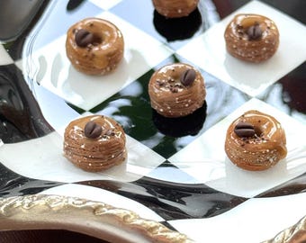 Peut inclure: Six mini-pâtisseries, chacune garnie d'un grain de café, sont disposées sur une assiette à carreaux noirs et blancs. Les pâtisseries sont brun clair et glacées. L'assiette a une bordure dorée.