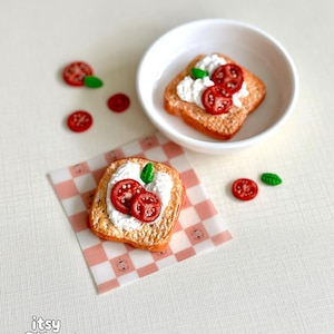 May include: Miniature food replicas featuring two pieces of toast topped with white burrata cheese, sliced red tomatoes, and green basil leaves. One toast sits in a small white bowl, while the other rests on a pink and white checkered paper square.