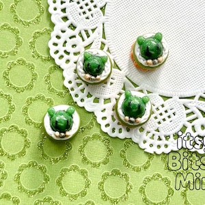 May include: Four handmade frog-shaped polymer clay earrings. Each earring features a green frog face on a white base, surrounded by a light green and gold border. The earrings are set against a green background with a white lace doily.