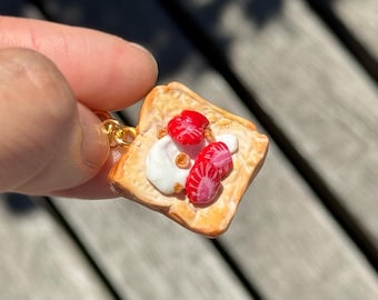 May include: A miniature toast-shaped keychain charm, featuring white cream and red strawberry slices. The charm is attached to a gold-coloured chain. The toast is a square shape.
