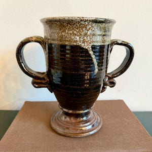 May include: A handcrafted ceramic mug with two handles. The mug features a dark brown base with a lighter brown speckled rim and a pedestal base. The interior is a lighter brown color. The mug is sitting on a brown book.