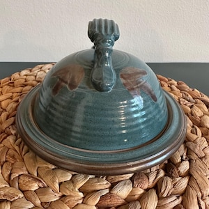 May include: A teal ceramic butter dish with a lid and handle. The dish has a round base and a domed lid with a textured handle. Brown leaf-like designs are painted on the lid. The dish sits on a woven placemat.