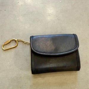 May include: A dark brown leather wallet with a fold-over flap and a gold-toned metal chain attached to a matching gold-toned carabiner clip. The wallet features a small embossed Coach logo on the bottom right corner of the front exterior surface.