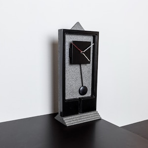 May include: A black and grey pendulum clock with a square face and a black frame. The clock is on a black surface.