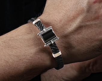 May include: A black leather bracelet with a square, black gemstone centerpiece and silver accents. The bracelet has a braided design and is worn on a wrist. A silver ring with a floral pattern is also visible.