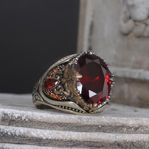 Könnte beinhalten: Ein silberner Ring mit einem großen, ovalen roten Edelstein, der von kleineren roten Edelsteinen umgeben ist. Der Ring ist vor einem weißen, strukturierten Hintergrund platziert.