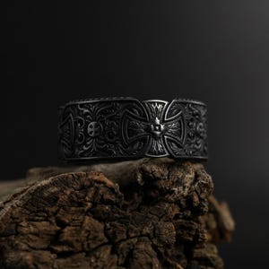 May include: A dark grey metal cuff bracelet with an ornate, engraved design. The bracelet features a central cross motif and detailed floral and skull patterns. The bracelet is displayed on a piece of weathered wood against a dark background.