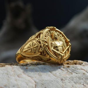 Puede incluir: Un anillo dorado con un dise&ntilde;o intrincado que presenta las palabras "San Miguel" y una imagen de un &aacute;ngel sosteniendo una espada. El anillo est&aacute; sobre una piedra de color claro, mostrando su detallada artesan&iacute;a.