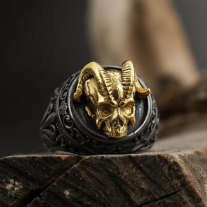 Può includere: Un anello argentato con un design a teschio dorato e corna di ariete. Il teschio è centrato su una fascia scura e decorata. L'anello è esposto su legno, mettendo in risalto i suoi dettagli intricati e lo stile gotico.