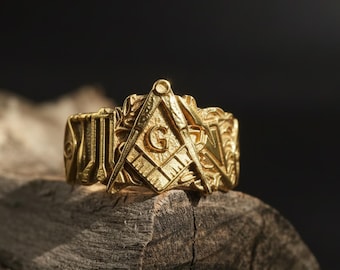 May include: A gold-colored ring featuring Masonic symbols, including a square and compass with the letter "G" in the center. The ring is highly detailed with intricate patterns and sits on a piece of weathered wood.
