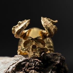 Può includere: Un anello color oro con un design a teschio con corna prominenti. L'anello è molto dettagliato, con tratti del viso visibili e corna strutturate. L'anello è esposto su un pezzo di legno su sfondo scuro.