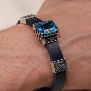 May include: A blue leather bracelet with a large, square, light blue gemstone at the center. The bracelet has silver-colored metal accents and is secured with a clasp. The bracelet is worn on a wrist.