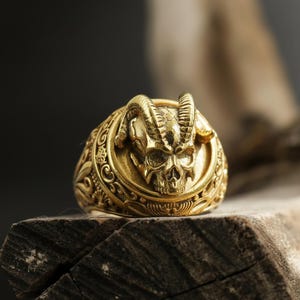 Può includere: Un anello dorato con un design dettagliato a teschio, con corna e motivi intricati. L'anello è posto su una superficie di legno, mostrando la sua lavorazione ornata. Questo anello a teschio è un pezzo audace.