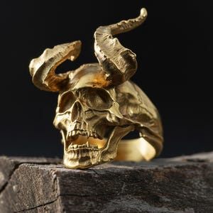 Può includere: Un anello color oro con un design dettagliato a teschio con corna ricurve. L'anello è posto su uno sfondo scuro, evidenziando gli intricati dettagli del teschio e la consistenza delle corna. L'anello è posizionato su una superficie di legno.