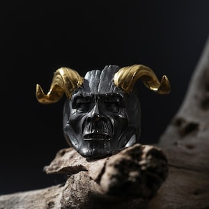 May include: A silver-toned ring featuring a detailed demonic face with gold-colored horns. The ring has a textured surface and a dark, aged appearance. The design is a bold statement piece.