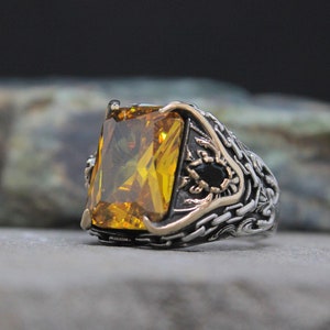May include: A silver ring with an ornate design featuring a large, rectangular, yellow gemstone. The ring has a black stone accent.