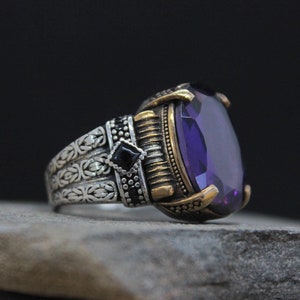 May include: A silver and gold ring with a large purple gemstone and smaller black gemstones. The ring is on a gray rock.