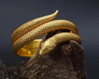 May include: A gold-colored ring in the shape of a snake, coiled around a finger. The snake's head is visible, with detailed scales and a textured surface. The ring is displayed on a piece of dark, textured wood against a dark background.
