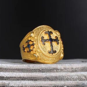 Könnte beinhalten: Ein goldfarbener Ring mit Kreuzmuster. Der Ring hat ein zentrales Kreuz mit einem blauen Edelstein und einen strukturierten Hintergrund. Der Ring wird auf einer Steinoberfläche präsentiert.