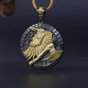 May include: A gold and silver pendant necklace featuring a detailed raven with outstretched wings and an axe. The raven is gold, set against a silver background, and hangs from a gold chain.