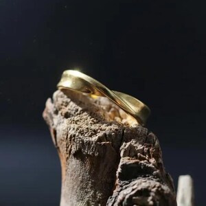 May include: A gold ring with a smooth, twisted, continuous band design. The ring features a polished metallic finish that reflects light, creating a subtle sheen across its curved surface. The band has a uniform width and a rounded, flowing shape.