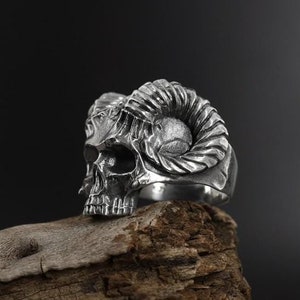 Può includere: Un anello d'argento a forma di teschio con corna di ariete. L'anello è appoggiato su un pezzo di legno consumato.