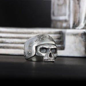 Könnte beinhalten: Silberfarbener Totenkopfring mit Motorradhelm-Design. Auf dem Helm steht "Ride Lite". Der Ring ist mit Nieten und einem Totenkopfgesicht versehen.
