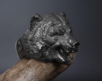 May include: A silver ring with a detailed bear head design. The bear's mouth is open and its teeth are visible. The ring is worn on a finger.