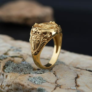 Puede incluir: Un anillo dorado con un dise&ntilde;o ornamentado. El anillo presenta un escudo con una cruz, rodeado de intrincados patrones florales. El anillo est&aacute; sobre una superficie de piedra natural.
