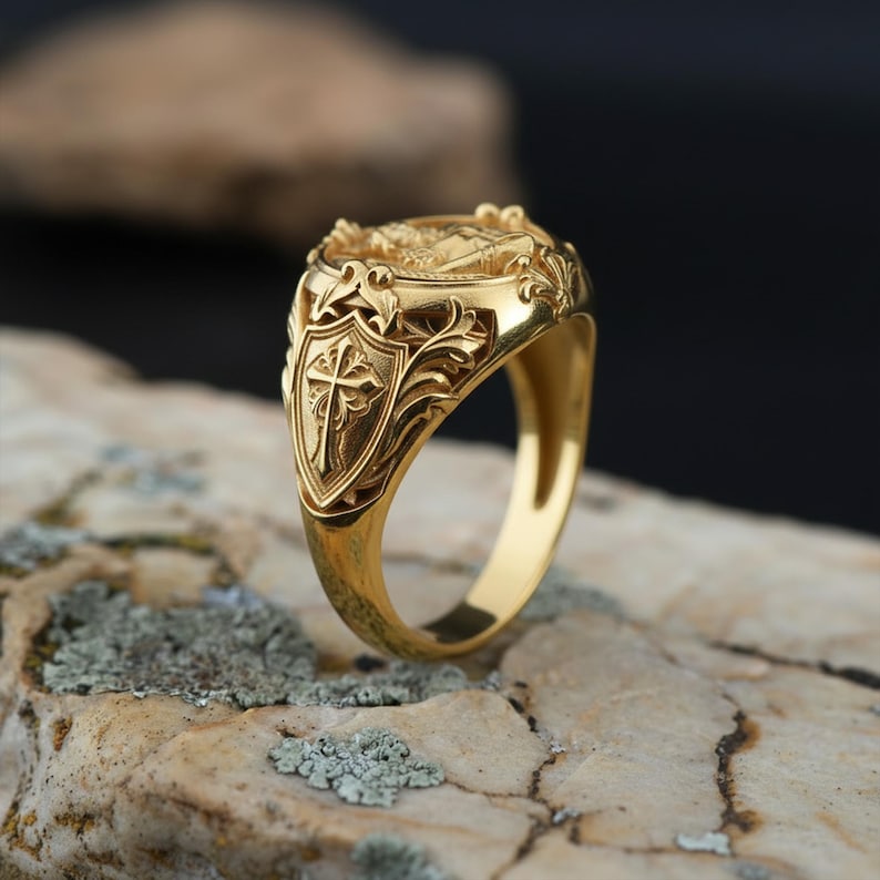 Puede incluir: Un anillo dorado con un dise&ntilde;o ornamentado. El anillo presenta un escudo con una cruz, rodeado de intrincados patrones florales. El anillo est&aacute; sobre una superficie de piedra natural.