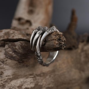 Op de afbeelding: Een zilveren ring met een gedetailleerd klauwontwerp. De ring is gemaakt van metaal en heeft een textuur.