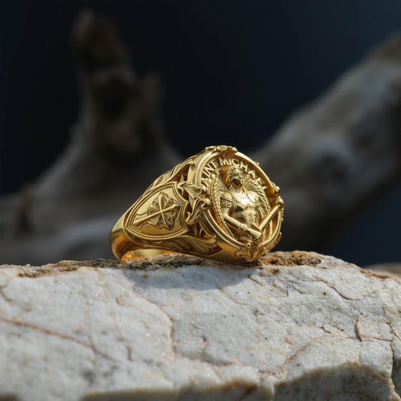 Puede incluir: Un anillo dorado con un dise&ntilde;o intrincado que presenta las palabras "San Miguel" y una imagen de un &aacute;ngel sosteniendo una espada. El anillo est&aacute; sobre una piedra de color claro, mostrando su detallada artesan&iacute;a.