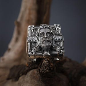 May include: A silver ring with a detailed design featuring a face of Jesus Christ. The ring is square shaped and has a cross design.