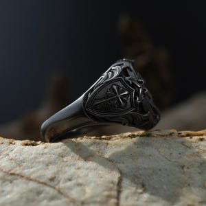 Puede incluir: Un anillo de sello gris oscuro con un dise&ntilde;o de escudo ornamentado con una cruz y detalles florales. El anillo est&aacute; colocado sobre una superficie clara y texturizada, mostrando los intrincados detalles del trabajo en metal.