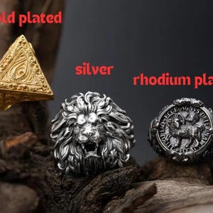 Puede incluir: Tres anillos: un anillo piramidal chapado en oro, un anillo de le&oacute;n plateado y un anillo rodiado con un dise&ntilde;o de cordero. Los anillos se exhiben sobre una superficie oscura y texturizada.