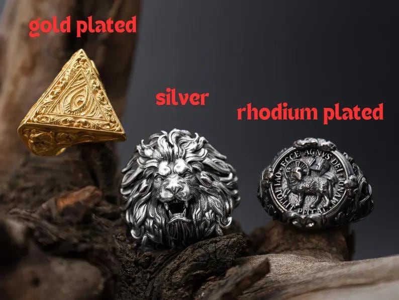 Puede incluir: Tres anillos: un anillo piramidal chapado en oro, un anillo de le&oacute;n plateado y un anillo rodiado con un dise&ntilde;o de cordero. Los anillos se exhiben sobre una superficie oscura y texturizada.