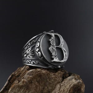 Op de afbeelding: Een zilveren ring met een sierlijk ontwerp met de letter "B" in het midden. De ring ligt op een stuk hout.