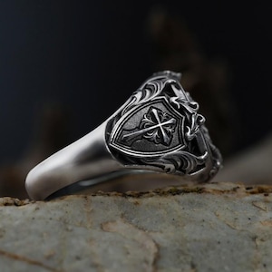 Puede incluir: Un anillo de plata con un dise&ntilde;o ornamentado. El anillo presenta un escudo con una cruz en el centro, rodeado de detalles de volutas. El anillo se encuentra sobre un fondo borroso, resaltando los intrincados detalles de la joya.