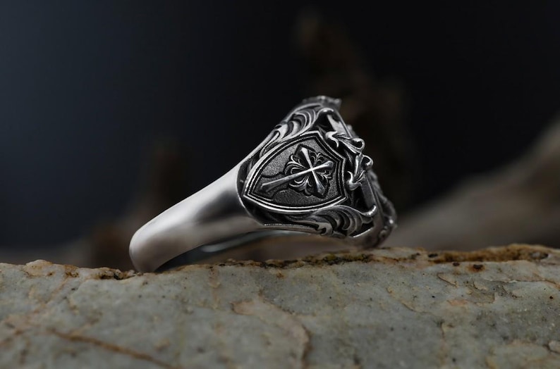 Puede incluir: Un anillo de plata con un dise&ntilde;o ornamentado. El anillo presenta un escudo con una cruz en el centro, rodeado de detalles de volutas. El anillo se encuentra sobre un fondo borroso, resaltando los intrincados detalles de la joya.