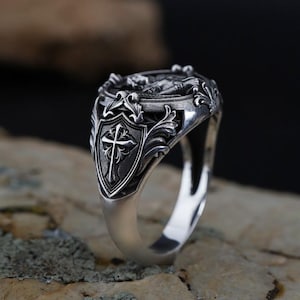 Puede incluir: Un anillo plateado con un dise&ntilde;o ornamentado. El anillo presenta un escudo con una cruz y detalles de volutas. La parte superior del anillo tiene un dise&ntilde;o en relieve detallado. El anillo est&aacute; sobre un fondo neutro.