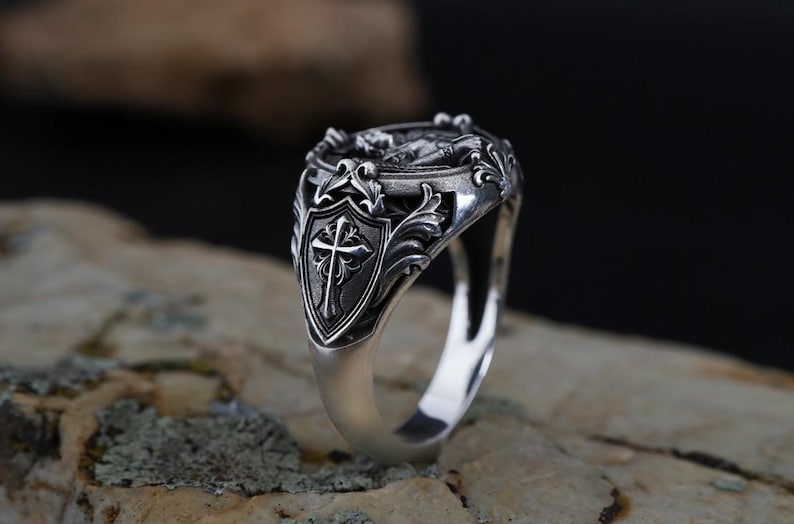 Puede incluir: Un anillo plateado con un dise&ntilde;o ornamentado. El anillo presenta un escudo con una cruz y detalles de volutas. La parte superior del anillo tiene un dise&ntilde;o en relieve detallado. El anillo est&aacute; sobre un fondo neutro.
