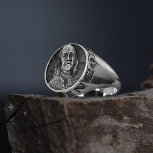 Könnte beinhalten: Ein silberner Ring mit einem erhabenen Bild von Benjamin Franklin auf der Vorderseite. Der Ring liegt auf einem Stück Holz.