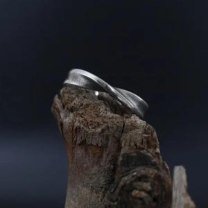 May include: A silver ring with a twisted design, resting on a piece of weathered wood.