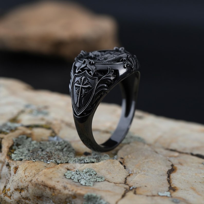 Puede incluir: Un anillo de sello negro con un dise&ntilde;o de escudo con una cruz. El anillo tiene intrincados detalles florales y decorativos. El anillo se muestra sobre una superficie clara y texturizada.