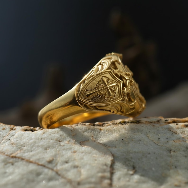 Puede incluir: Un anillo dorado con un dise&ntilde;o ornamentado. El anillo presenta un emblema en forma de escudo con una cruz y intrincados patrones florales. El anillo est&aacute; sobre una superficie clara y texturizada.